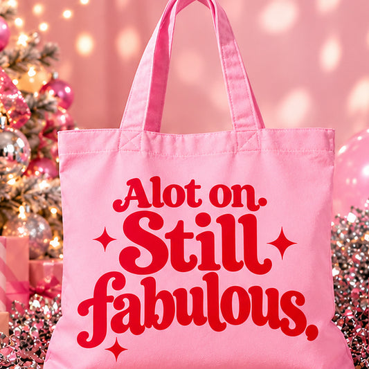 Pink organic cotton tote bag with red retro “A lot on. Still fabulous.” lettering, photographed with festive pink decorations and silver disco balls.