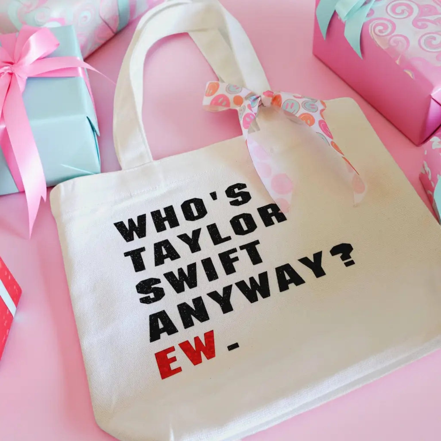 White canvas tote bag with bold black text reading 'WHO'S TAYLOR SWIFT ANYWAY? EW.' featuring a colorful smiley face ribbon tied to the handle - Boutique Swiftie party favors by Minty and Mallow.